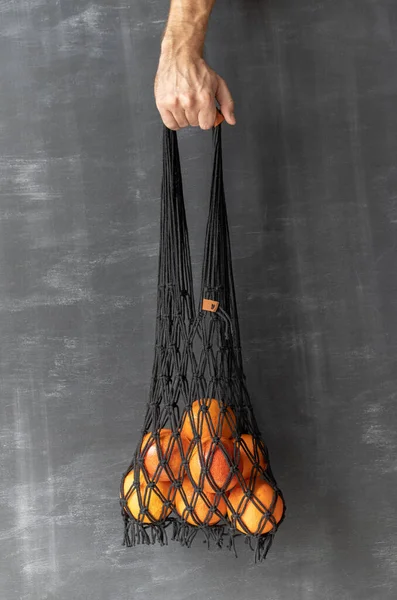 A mans hand holds a string bag in a macrame, tied with a black rope, with tangerines and oranges on a dark background of a chalky wall.