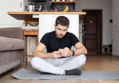 Spor giyimli bir adam apartmanın içinde yoga minderinde oturmuş akıllı telefondan online yayın izleyerek ders çalışıyor. Ev zindeliği