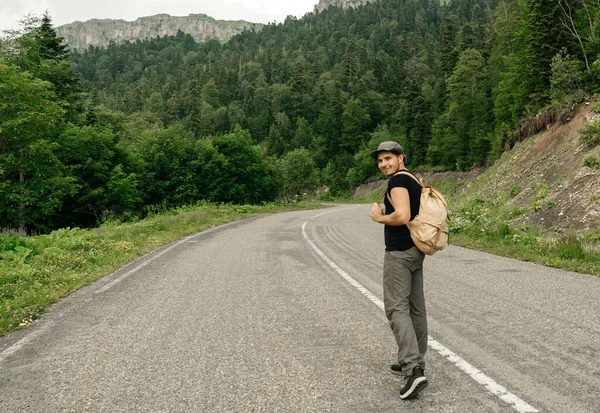 traveler man hitchhiking, standing on the road with a backpack in a hat on a background of a mountain landscape.