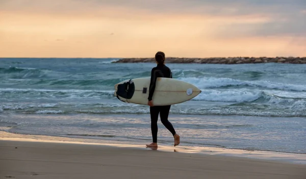 La chica deportiva va a surfear. Mujer en traje de neopreno y puesta de sol o sunris — Foto de Stock