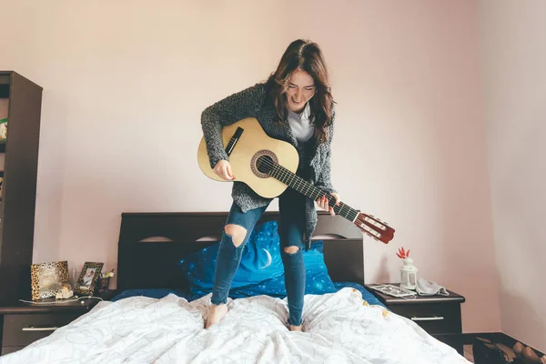 年轻美丽的东方女人站在床上弹奏吉它 女孩的力量 有乐趣的概念 — 图库照片