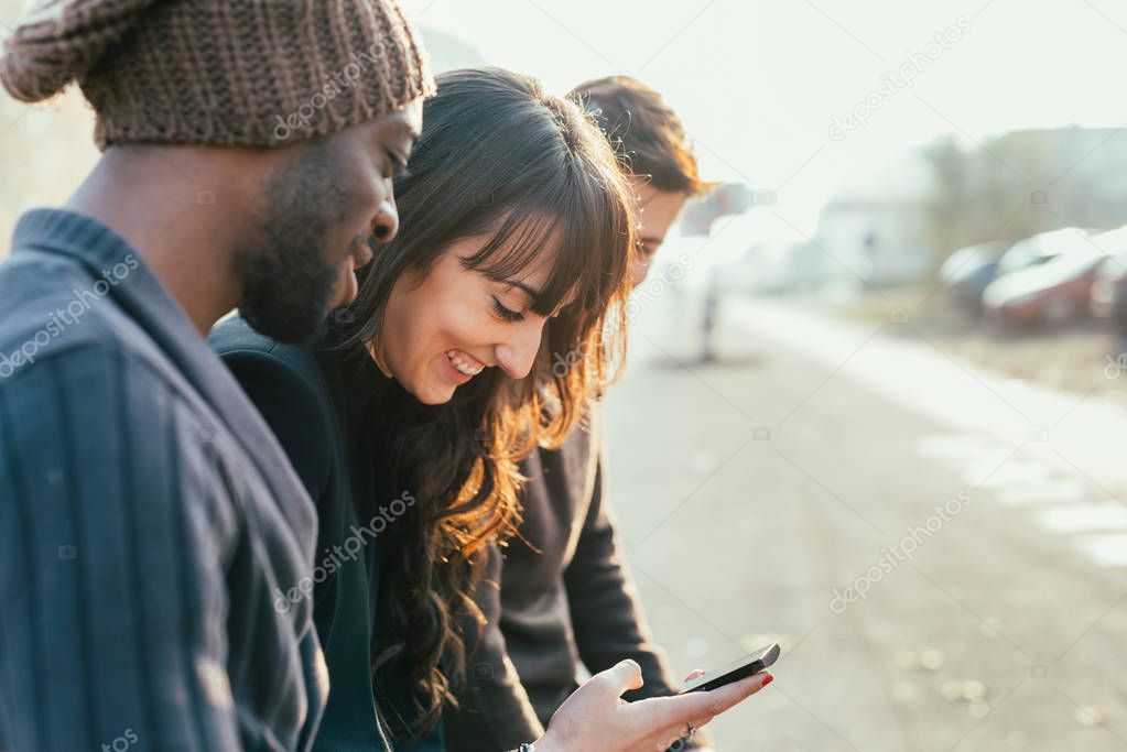Three multiethnic woman and men friends outdoor in city back light, chatting and holding smart phone - friendship, social network, concept 