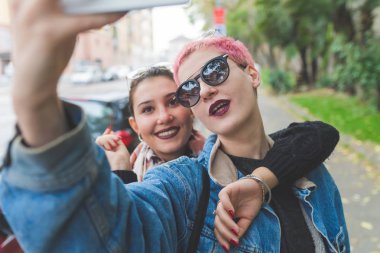 İki genç kadın akıllı telefon kullanarak selfie çekiyor. Teknoloji, sosyal ağ, gençlik kültürü.
