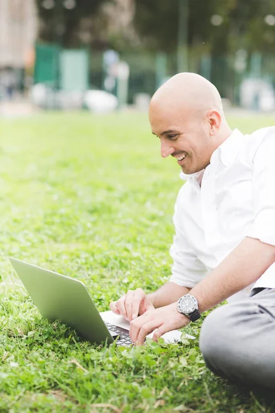 Young contemporary businessman remote working lying down outdoor in a city park using computer