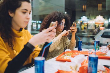 İki genç kadın fast food 'da akıllı telefon kullanarak yemek yiyor. Yiyecek, içecek, müşteri, sosyal toplanma kavramı.