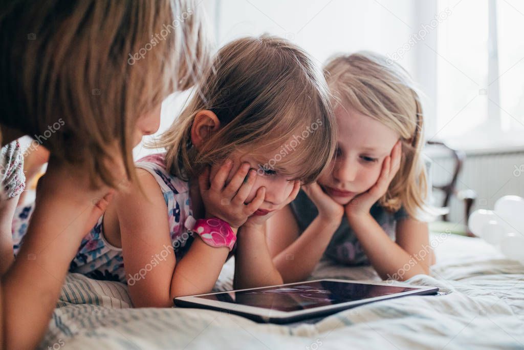 Three little sisters indoor at home using tablet lying in bed