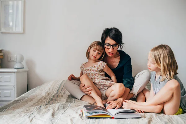 Mother of two female children sitting on bed reading book together — 스톡 사진