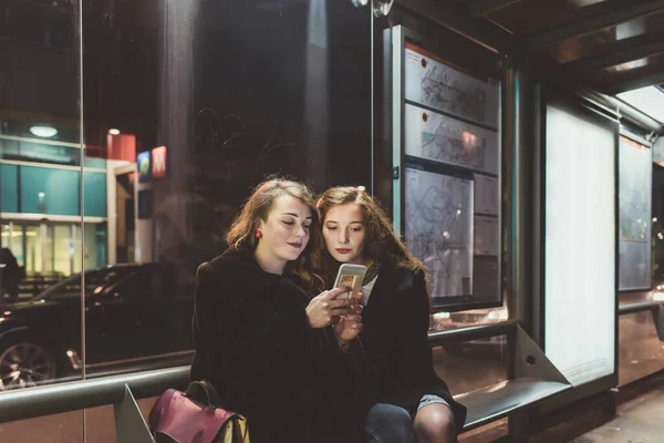 Δύο Νεαρές Όμορφες Γυναίκες Κάθονται Στάση Λεωφορείου Χρησιμοποιώντας Smartphone Αναμονή — Φωτογραφία Αρχείου