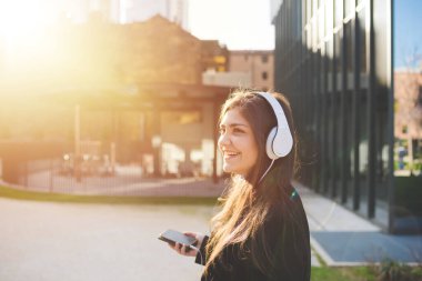 Genç, güzel, güler yüzlü, dışarıda mutlu bir kadın akıllı telefon kullanıyor, müzik dinliyor. Akış, mutluluk, keyif konsepti.