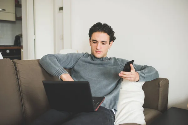 若い美しい白人男性屋内の自宅で横にソファを使用してコンピュータとスマートフォン リモート作業 ストリーミングテレビ フリーランスのコンセプト — ストック写真