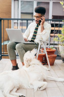 Terasta bilgisayarla konuşan akıllı telefon kullanan yakışıklı bir zenci. Ev işi, çoklu görev, iş konsepti.