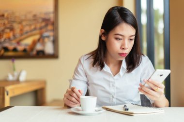 Bir kahve fincanı ve defter ile akıllı telefon kullanan genç kadın.