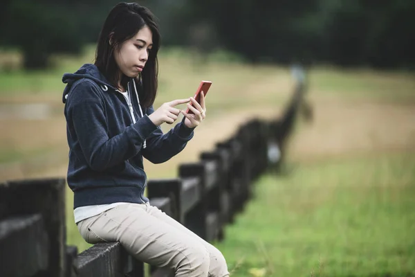 Wanita muda yang bahagia duduk di pagar dan menggunakan smartphone-nya untuk — Stok Foto