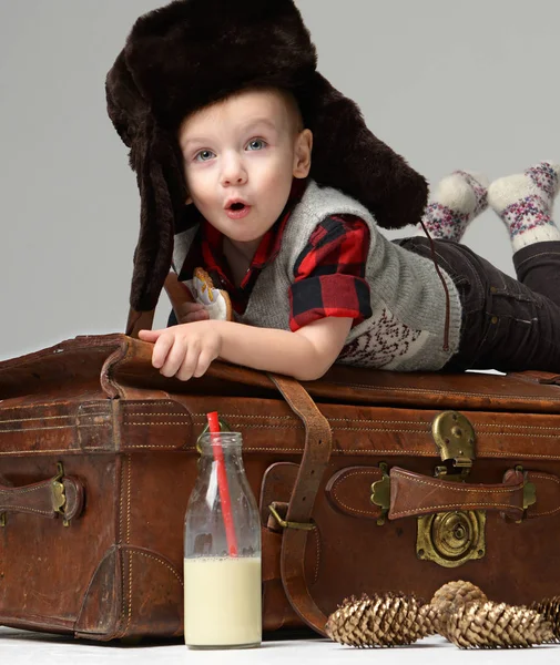 Small child baby boy in winter hat lying on retro leather travel bag in winter snow hat  with bottle of milk — Stock Photo, Image