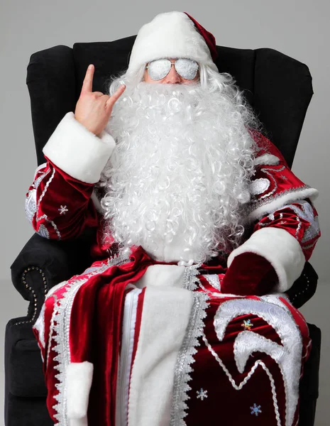 Bad santa claus in snow flakes sunglasses sitting and showing rock sign — Stock Photo, Image