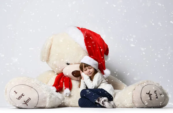 Junges Mädchen sitzt und schläft mit großen weichen Teddybär-Spielzeug in Weihnachtsmann rote Mütze unter schwerem Schnee — Stockfoto