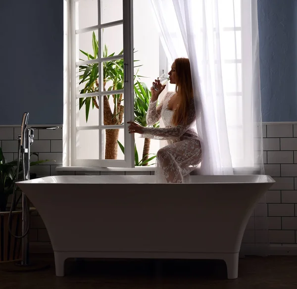 Wanita cantik muda minum air dekat bak mandi dan membuka jendela kamar mandi — Stok Foto