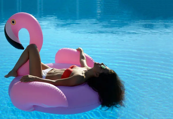 在一个巨大的充气巨型粉红色火烈鸟浮动床垫在泳池的女人在红色比基尼 — 图库照片