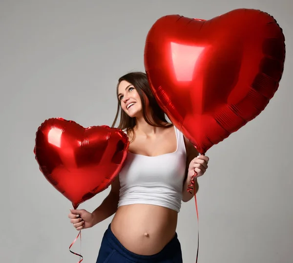 Junge schöne schwangere Frau mit roten Herzballons zum Valentinstag glücklich lächelnd — Stockfoto