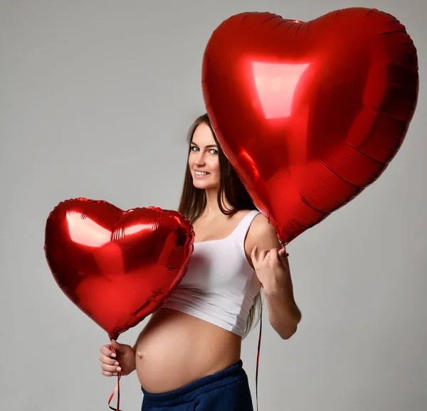 Joven mujer embarazada hermosa con globos de corazón rojo para el día de San Valentín feliz sonrisa —  Fotos de Stock