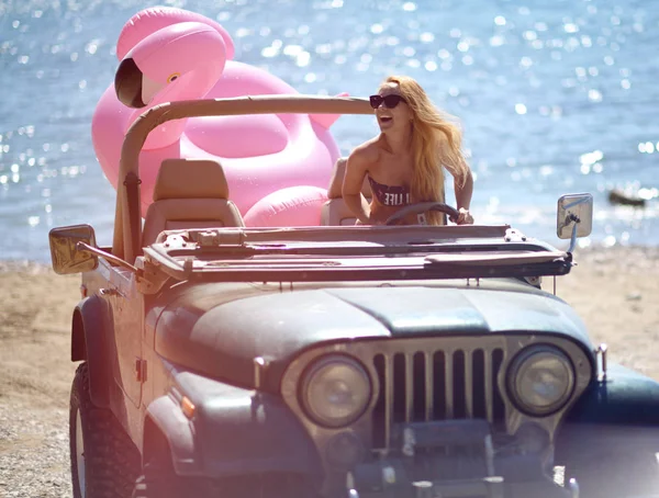 Jovem menina em óculos de sol sentado no carro caminhão de praia no dia ensolarado de verão perto da praia do mar com gigante inflável gigante rosa flamingo flutuador colchão — Fotografia de Stock