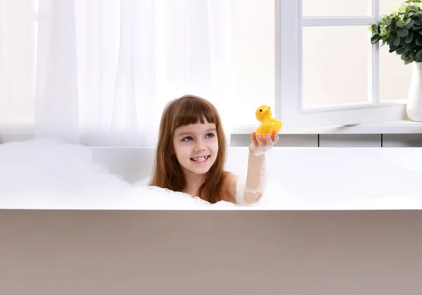 Feliz niña sentada en la bañera jugando con el juguete del pato amarillo en el baño —  Fotos de Stock