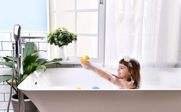Feliz menina sentada na banheira brincando com brinquedos de pato no banheiro — Fotografia de Stock