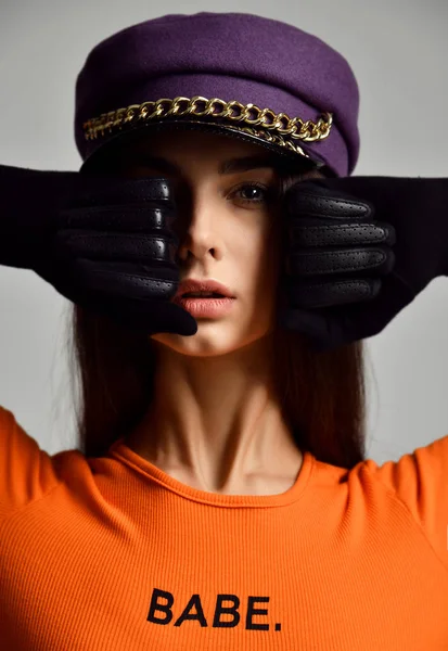 Potret wanita muda berambut cokelat seksi dengan topi puncak ungu atau baret dengan rantai emas dengan sarung tangan kulit dan kemeja oranye — Stok Foto
