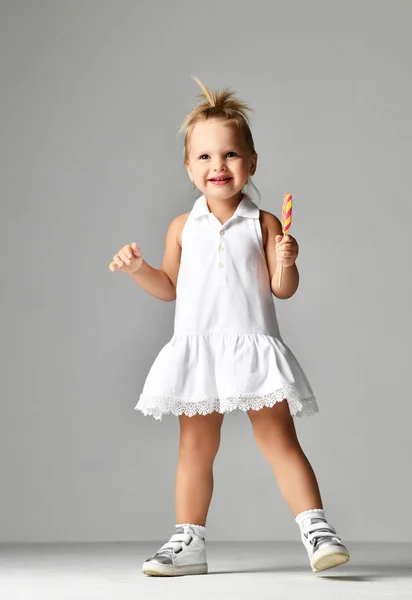 Молодая красивая малышка девочка с большой сладкой конфеткой леденец в белом платье на серой — стоковое фото