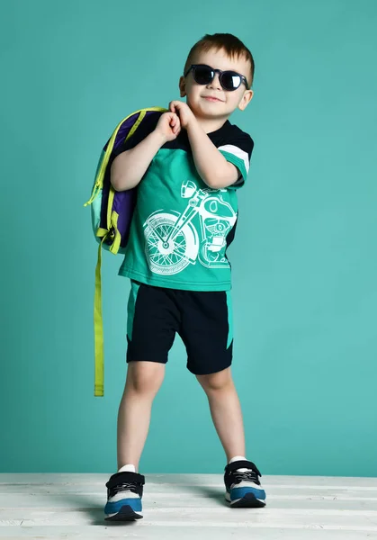 Niño preescolar de pie con mochila y gafas de sol en pantalones cortos y camiseta en menta verde —  Fotos de Stock