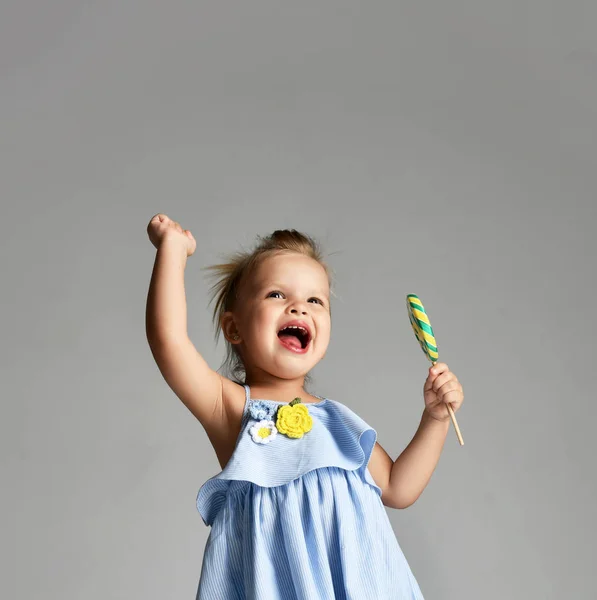 Молодая красивая малышка девочка с большой сладкой конфеткой леденец в синем платье на сером — стоковое фото
