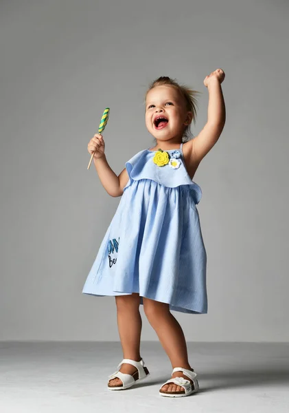 Young pretty toddler girl kid with big  sweet lollypop candy in blue dress on grey — Stock Photo, Image