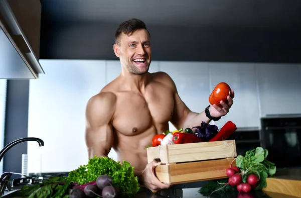 Muscular sport man standing on kitchen with groceries vegetables hold radish in hands looking at the corner smiling — Stock Photo, Image