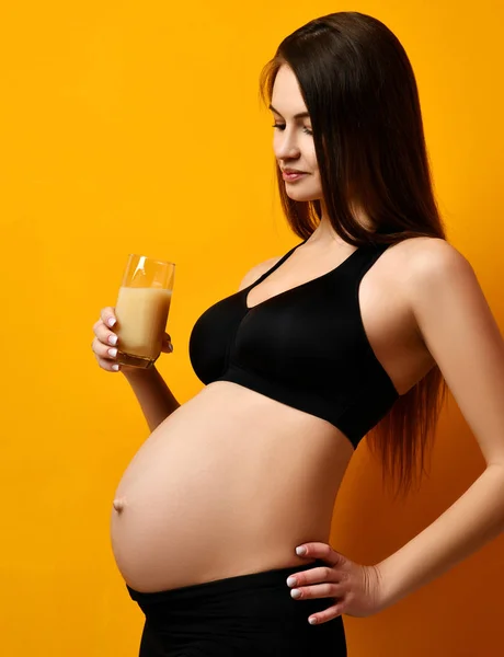 Беременная женщина в черной рубашке держит здоровый апельсиновый ананасовый сок улыбаясь на желтый — стоковое фото