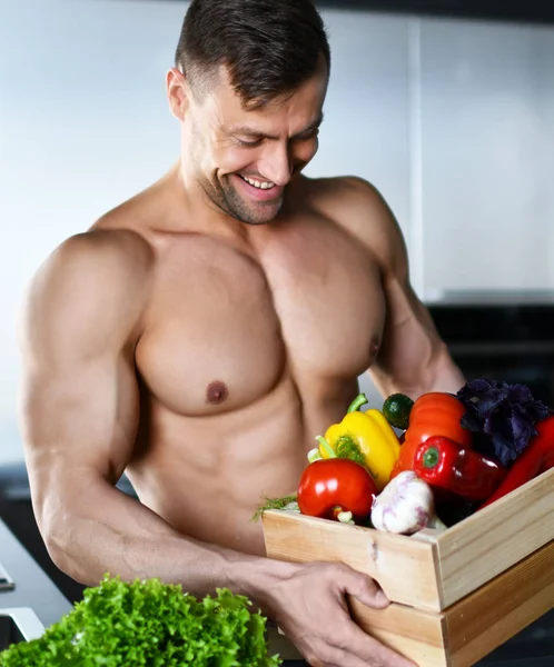 Strong muscular sport man standing on kitchen box full of fresh groceries vegetables in hands looking smiling — Stock Photo, Image