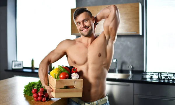 Strong muscular sport man standing on kitchen box full of fresh groceries vegetables in hands looking smiling — Stock Photo, Image