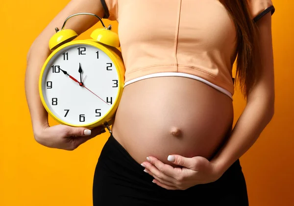 Zwangere vrouw in top shirt houden grote klok horloge glimlachend op geel — Stockfoto