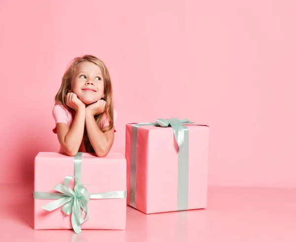 Navidad y Año Nuevo. Niza niña sentada detrás de cajas de regalos con la barbilla en las manos soñando, esperando en rosa — Foto de Stock