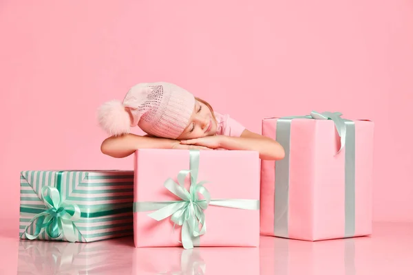 Koncept Vánoc a Nového roku. Roztomilá holčička v teplém klobouku spí, dřímá, sní o velkých dárkových krabicích na růžové — Stock fotografie