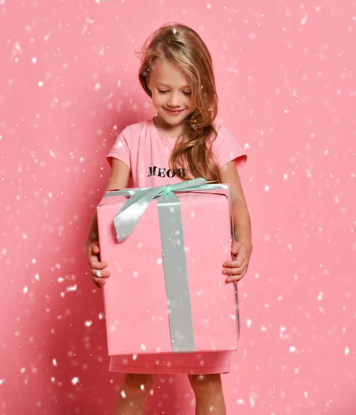 Joyful blonde kid girl in pink dress holding a big present gifts box looking at it impatiently on pink under the snow — Stock Photo, Image