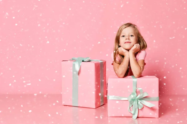 Christmas and New Year concept. Kid girl is sitting behind gifts with her chin in hands under the snow on pink — Stock Photo, Image