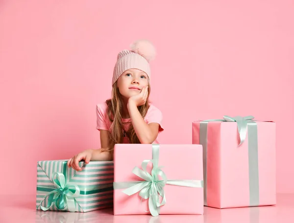 Pensive kid girl is sitting behind gifts boxes presents guessing, dreaming of a gift, waiting on pink — Stock fotografie