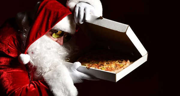 Papai Noel em óculos de sol levantando a tampa da caixa de pizza e olhando para dentro, assistindo, desejando. Ano Novo e Xmas fast food — Fotografia de Stock