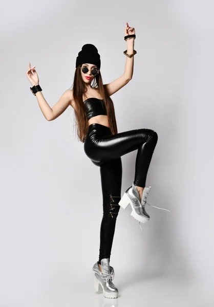 Brutal ung kvinna i svart hatt, tight läder leggings med blixtlås och runda solglasögon dansar med händerna upp — Stockfoto