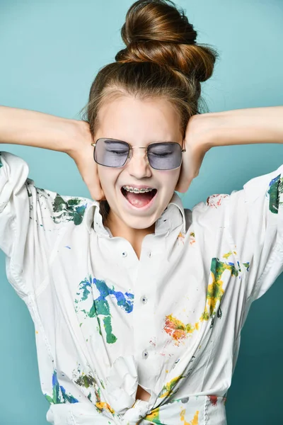 Retrato de menina com aparelho em camisa branca com manchas de tinta e óculos cobrindo os ouvidos com as mãos — Fotografia de Stock