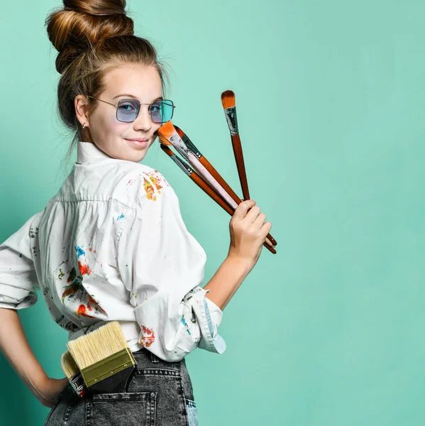 Sonriente joven adolescente en camisa blanca con manchas de pintura con un conjunto de pinceles nos mira hacia atrás, listo para dibujar — Foto de Stock