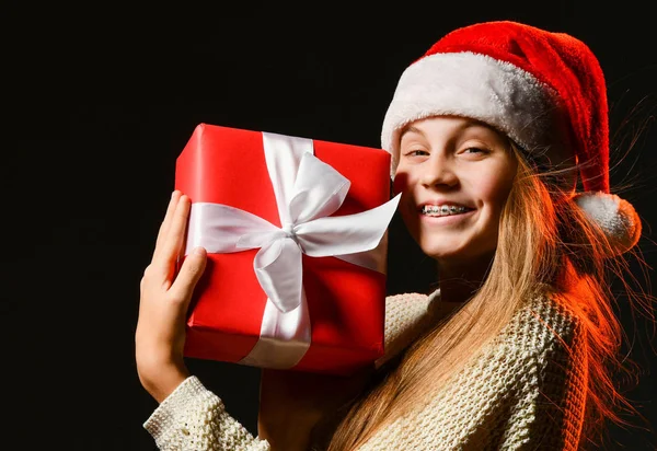 Glad leende tonåring flicka i röd mössa med pompon med hängslen håller helong efterlängtade nyår gåva röd låda med band — Stockfoto