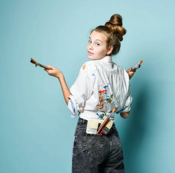 Glimlachende jonge kunstenaar aan het werk in hemd met verf vlekken staat achterover en kijkt over haar schouder naar ons — Stockfoto