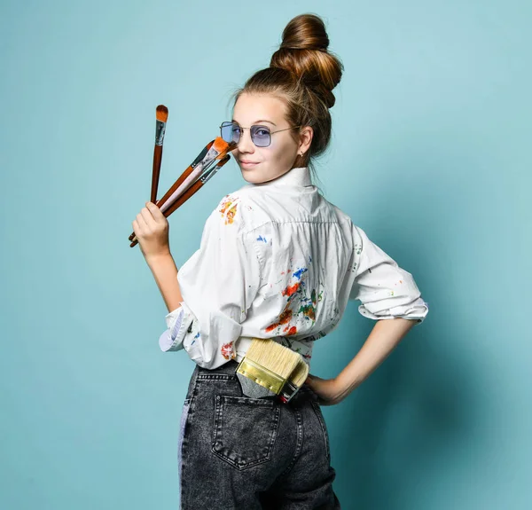 Jovem artista mulher com um conjunto de pincéis em camisa com manchas de tinta está no trabalho, de pé, olhando por cima de seu ombro — Fotografia de Stock