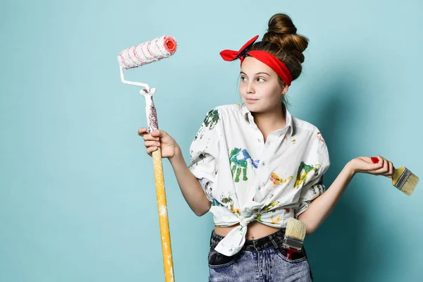 Jovem artista pintor de camisa com manchas de tinta está segurando pincel e rolo, olhando para o lado com uma ligeira surpresa — Fotografia de Stock